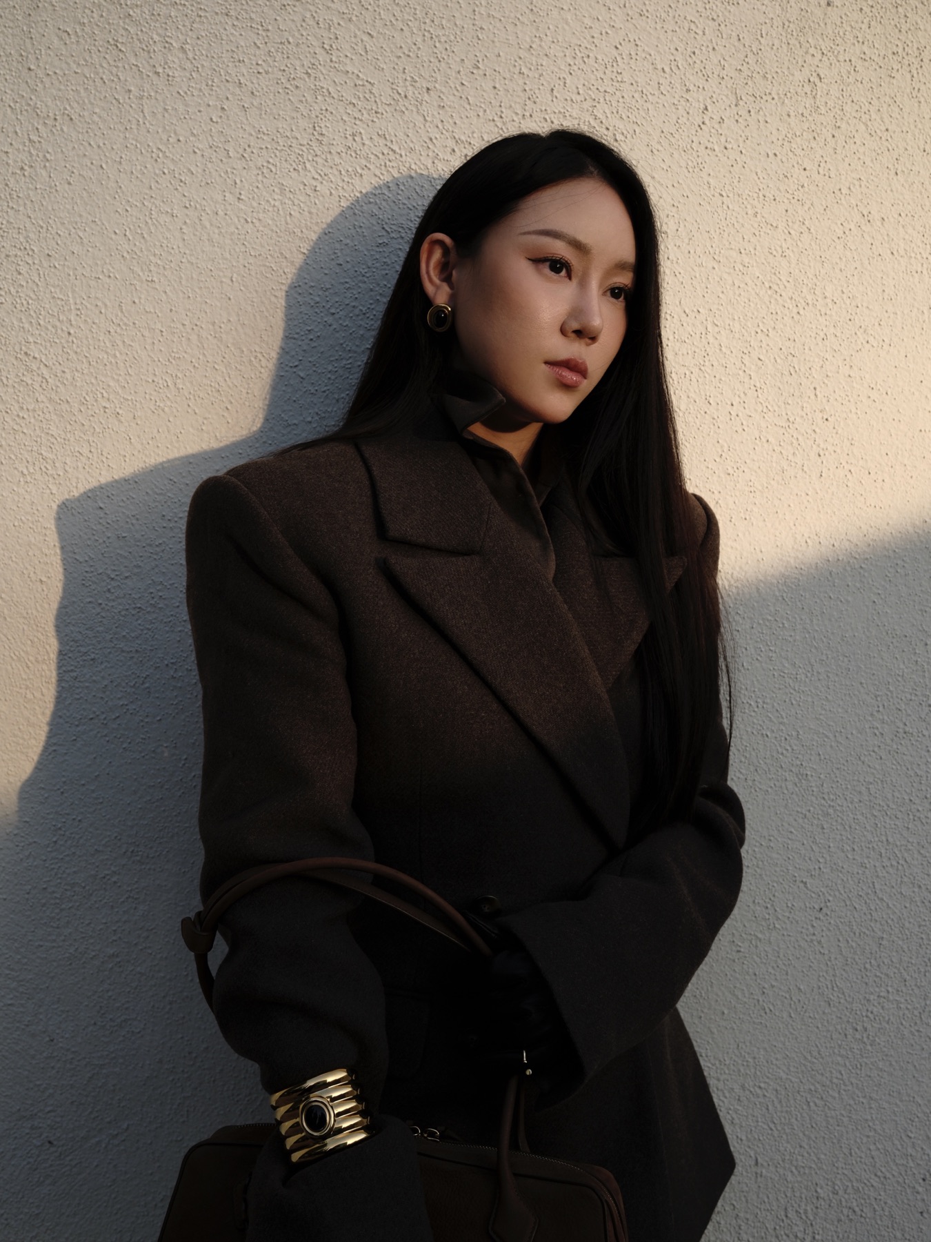 PHOEBE.L in dark tailoring against a sunlit textured wall